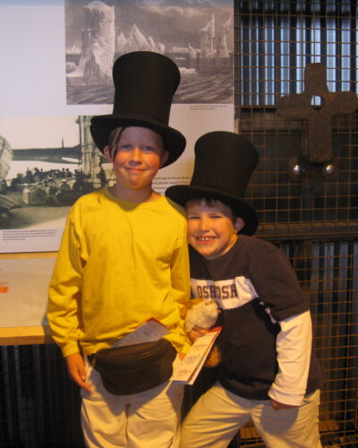 Two children wearing top hats, smiling indoors.