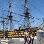 Historic warship docked with visitors nearby.