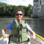 Person rowing boat near stone building, trees.