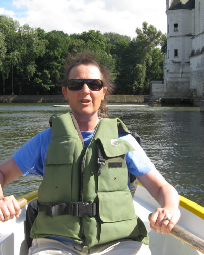 Person rowing boat near stone building, trees.