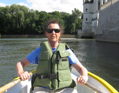 Person rowing boat near stone building, trees.
