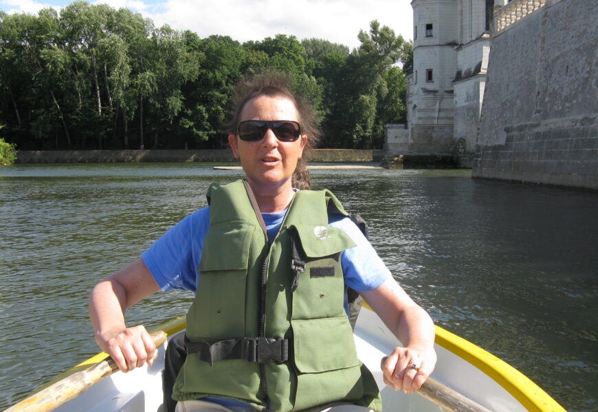 Person rowing boat near stone building, trees.